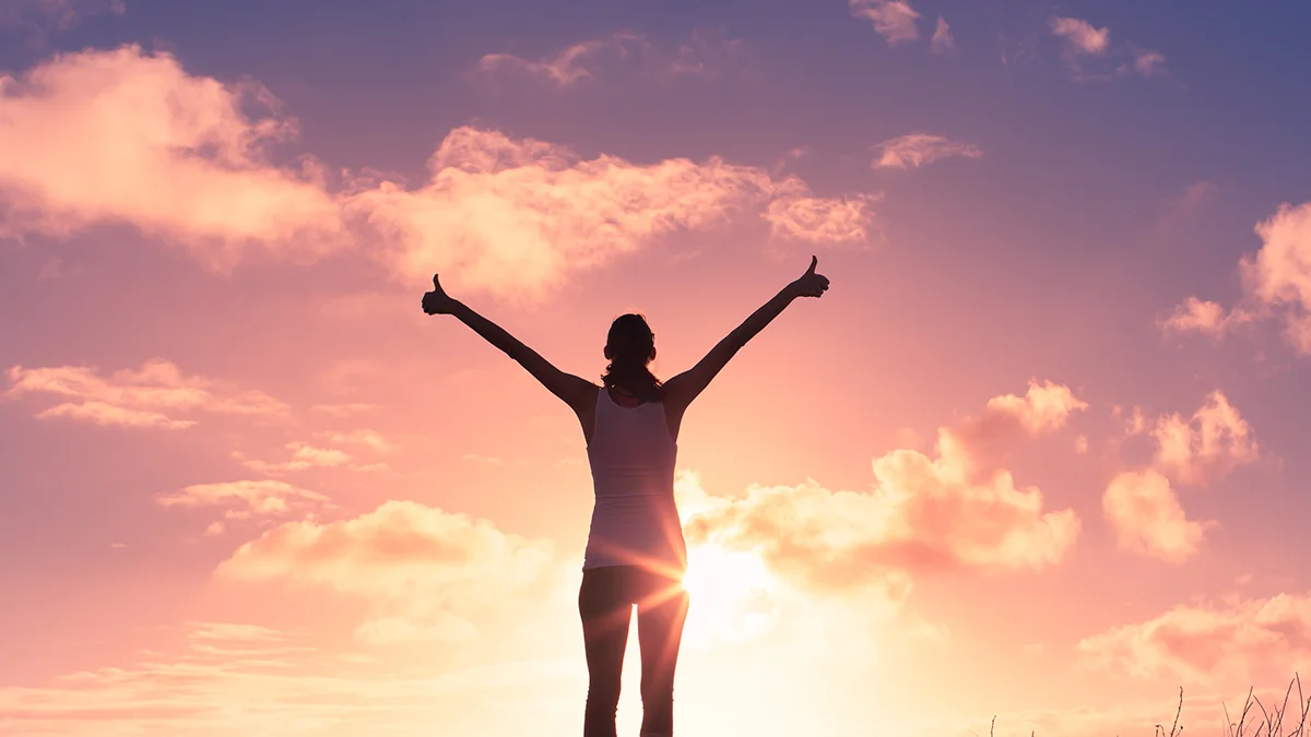 An outline of a woman standing tall with her hands towards the sky, watching a beautiful sunset